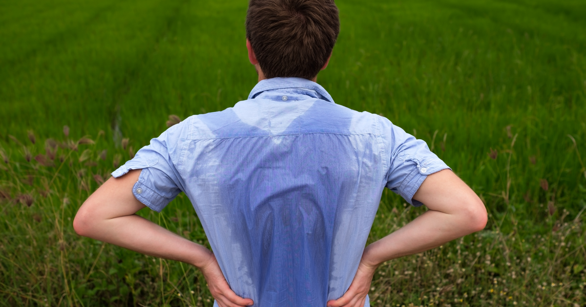 A man standing in an open green field with a sweat patch across the back of his blue shirt, illustrating excessive sweating in the Australian and New Zealand climate