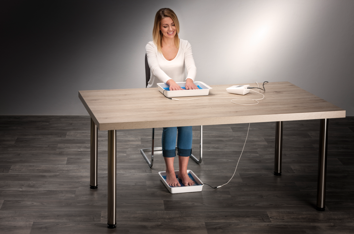 Woman seated at a table with hands and feet submerged in water trays connected to a Saalio iontophoresis device.
