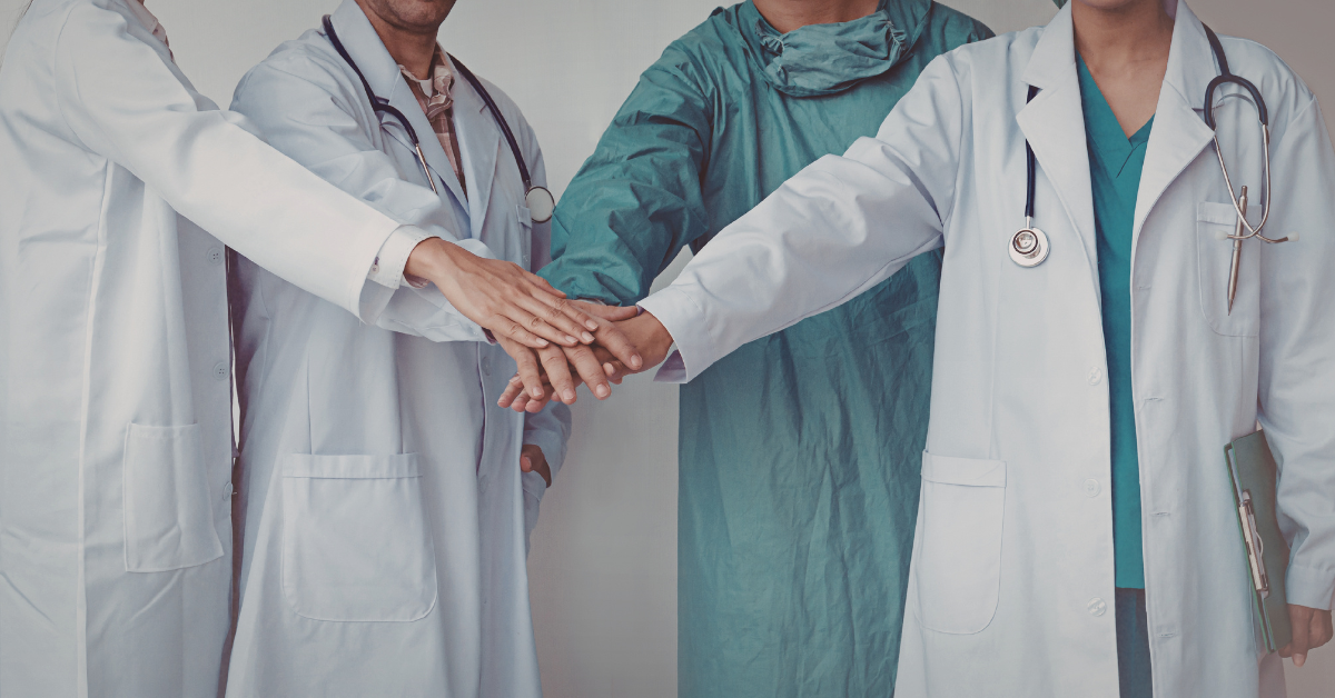 Three medical professionals in lab coats and scrubs joining hands together, representing collaborative healthcare and dermatologist-supported treatment approaches.